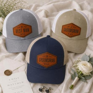 May include: Three baseball caps with mesh backs and leather-like patches. One grey cap reads BEST MAN, one tan cap reads GROOMSMAN, and one navy blue cap reads GROOMSMAN. Each patch includes initials and the date 06.14.26. The caps are arranged on a white fabric surface.