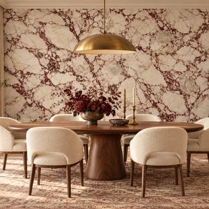 May include: A dining room featuring a large oval wooden table with a pedestal base, surrounded by six cream-colored upholstered chairs. A brass dome pendant light hangs above, and a wall with white and dark red marble pattern serves as a backdrop.