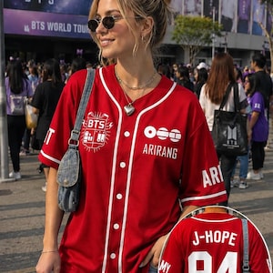 Op de afbeelding: Een rood honkbalshirt met korte mouwen, knoopsluiting en witte biezen. De voorkant is voorzien van een wit BTS-logo, de tekst ARIRANG en drie witte cirkels. De achterkant toont de witte tekst J-HOPE boven het grote witte nummer 94.
