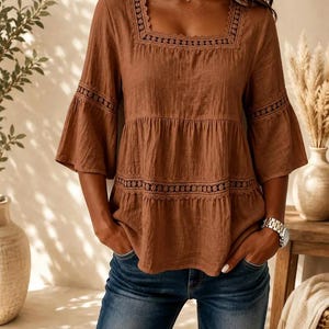 May include: A brown tiered blouse featuring a square neckline, three-quarter length bell sleeves, and intricate lace trim detailing along the neckline and horizontal tiers. The blouse is paired with blue denim jeans and a silver-toned wristwatch.