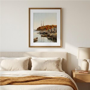 May include: A framed landscape painting depicting a harbor scene with several small boats moored near a rocky shoreline. The background features autumn foliage in shades of orange and red, with small wooden buildings situated along the water's edge.