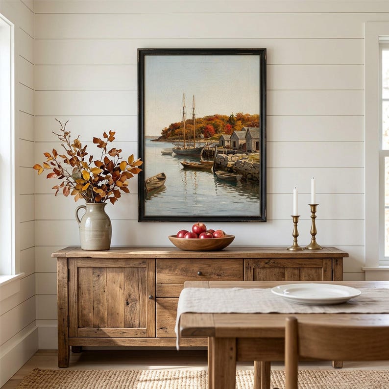 May include: A framed landscape painting depicting a harbor with sailboats and small wooden boats near a rocky shore with autumn foliage. The artwork is displayed above a wooden sideboard holding a ceramic pitcher with dried branches and a bowl of red apples.