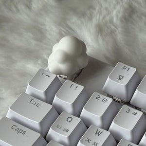以下が含まれることがあります： 白い雲の形をしたメカニカルキーボード用のキーキャップが、白いキーボードに取り付けられています。キーキャップは丸みを帯びた泡のような質感で、キーボードの左上部分に配置され、標準的なキーと置き換わっています。キーには黒い文字が印字されています。