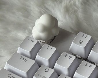May include: A white, cloud-shaped mechanical keyboard keycap sits on a white keyboard. The keycap has a rounded, bubbly texture and is positioned in the upper left area of the keyboard, replacing a standard key. The keys feature black lettering.