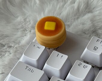 May include: A custom mechanical keyboard keycap shaped like a stack of two golden-brown pancakes topped with a square yellow pat of butter. The keycap is mounted on a white keyboard switch, positioned among white keys with black lettering.