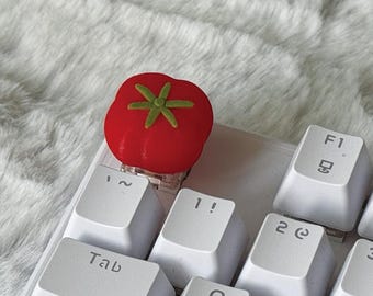 May include: A red tomato-shaped mechanical keyboard keycap with a green stem detail, positioned on a white keyboard above the Tab key. The keycap features a rounded, segmented design and is mounted on a clear switch base on the white keyboard.