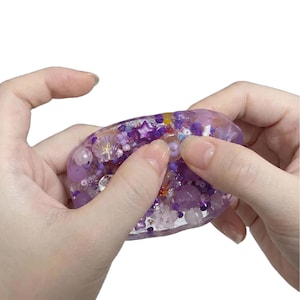 Puede incluir: Un juguete sensorial ovalado de resina púrpura translúcida relleno con una variedad de cuentas púrpuras y transparentes incrustadas, incluyendo elementos en forma de estrella, concha y esferas, sostenido entre dos manos sobre un fondo blanco.