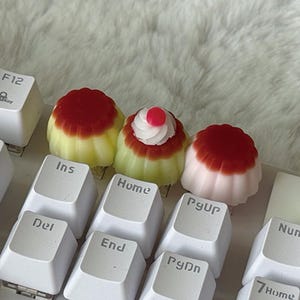 以下が含まれることがあります： ミニチュアデザートの形をした3つのカスタムメカニカルキーボード用キーキャップ。2つは赤いトップの黄色いプリンに似ており、1つは白いクリームの渦巻きと小さな赤い点が特徴で、白いキーボードに取り付けられています。