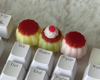 May include: Three custom mechanical keyboard keycaps shaped like miniature desserts. Two keycaps resemble yellow flan with red tops, and one features a white swirl of frosting with a small red dot. They are installed on a white keyboard.