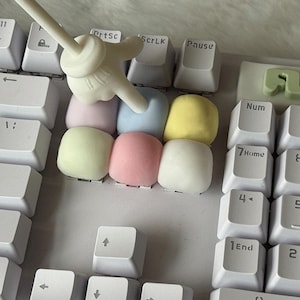 Peut inclure: Un clavier d'ordinateur blanc avec un groupe de six touches carrées aux couleurs pastel : rose clair, bleu, jaune et vert. Un outil de pointage blanc en forme de main de dessin animé appuie sur la touche bleu clair.