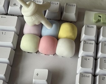 May include: A white computer keyboard featuring a cluster of six pastel-colored, square-shaped keycaps in light pink, blue, yellow, and green. A white, cartoon-style hand-shaped pointer tool is pressing down on the light blue keycap.