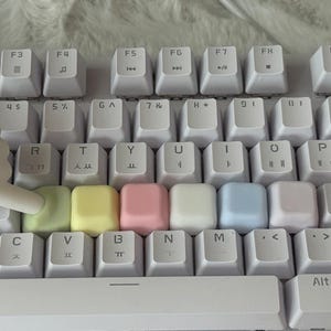 Könnte beinhalten: Eine weiße Computertastatur mit standardmäßigen alphanumerischen Tasten mit schwarzer Beschriftung und koreanischen Schriftzeichen. Eine Reihe von sechs leeren, pastellfarbenen Tastenkappen in Hellgrün, Gelb, Rosa, Weiß, Hellblau und Lavendel.