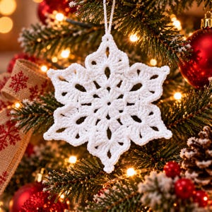 Puede incluir: Un adorno de copo de nieve tejido a ganchillo en color blanco con un diseño de estrella de seis puntas y patrones calados intrincados. El adorno cuenta con un fino lazo de hilo blanco en la parte superior para colgarlo.