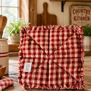 May include: A square quilted potholder featuring a red and white gingham check pattern with frayed edges. The fabric is stitched in a diagonal pattern meeting at the center. A small white rectangular label reads Sew Sunny in black text.