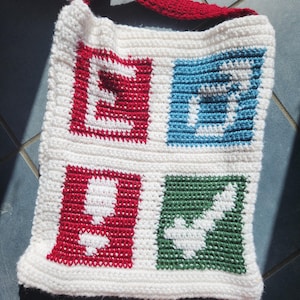 May include: A white crocheted tote bag featuring four square panels in a grid pattern. The top left panel is red with a white letter E, the top right is blue with a white letter S, the bottom left is red with a white exclamation mark, and the bottom right is green with a white checkmark.