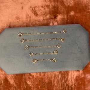 May include: Five gold-toned metal chain extenders of varying lengths, each featuring a circular spring ring clasp at both ends. The chains are arranged horizontally in descending order of length, showcasing a simple link design for jewelry.