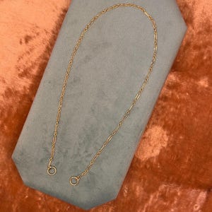 May include: A gold-toned chain necklace featuring a repeating pattern of elongated oval links connected by small circular links. The chain is arranged in a U-shape and terminates at both ends with a small, circular jump ring for attachment.