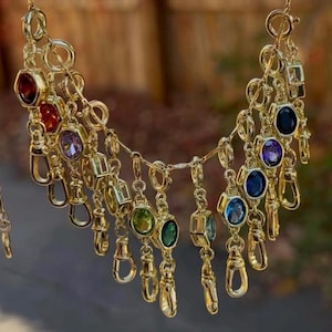 May include: A gold-toned chain necklace featuring multiple dangling charms, each consisting of a small gold lobster clasp attached to a faceted, bezel-set gemstone in various colors including red, purple, green, and blue.