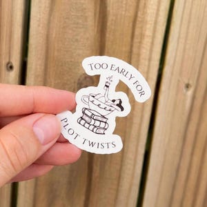 May include: A white die-cut sticker featuring a black line art illustration of a person relaxing in a teacup perched atop a stack of three books. The text "TOO EARLY FOR PLOT TWISTS" is printed in a black serif font, curving around the illustration.
