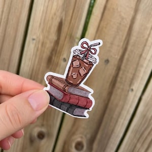 May include: A die-cut sticker featuring a stack of three books in shades of brown and red, topped with a clear plastic cup filled with iced coffee. The cup has a brown lid and a straw adorned with a small red bow, rendered in a flat illustration style.