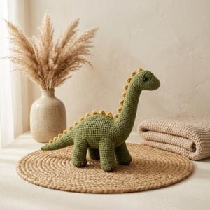 Puede incluir: Un juguete de dinosaurio tejido a mano en color verde oliva sólido. El dinosaurio presenta un cuello largo, un cuerpo redondeado y una hilera de pequeñas púas triangulares amarillas a lo largo de su espalda y cola. Tiene ojos oscuros y redondos.