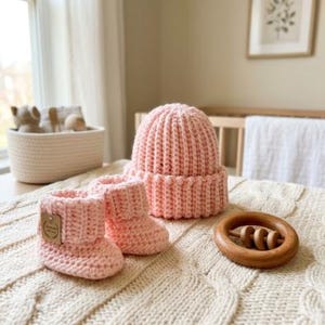 Puede incluir: Un gorro de ganchillo rosa claro con borde doblado y un par de patucos de ganchillo a juego en rosa claro con una pequeña etiqueta rectangular marrón en el lateral, junto a un anillo de dentición de madera con tres cuentas pequeñas.