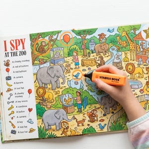 May include: An open I Spy At The Zoo activity book featuring colourful illustrations of zoo animals and a list of items to find. A hand holds an orange Stabilo Boss highlighter over the page, which shows elephants, monkeys, and a giraffe.