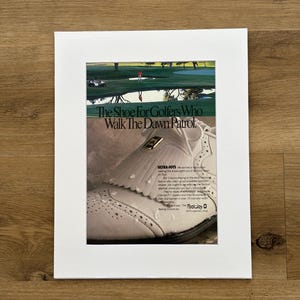 May include: A vintage advertisement print featuring a white golf shoe with a kiltie fringe and water droplets on the surface. The text reads The Shoe For Golfers Who Walk The Dawn Patrol and Ultra-Joys, with a golf course scene at the top.