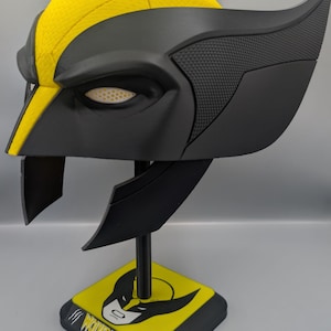 May include: A yellow and black Wolverine helmet featuring a textured finish, pointed side fins, and white mesh eye lenses. The helmet is mounted on a black display stand with a yellow base that displays a Wolverine graphic and the text WOLVERINE.