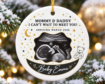 Può includere: Un ornamento rotondo in ceramica bianca con un'immagine ecografica in bianco e nero al centro. Il testo recita Mommy & Daddy I Can't Wait To Meet You! Arriving March 2026 e Baby Emma. È decorato con stelle dorate, una mezzaluna e un orsacchiotto.
