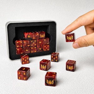 May include: A set of dark red and black marbled dice with gold-colored symbols. The dice feature teardrop and winged teardrop icons on their faces. Several dice are arranged inside a rectangular metal storage tin with black foam lining.