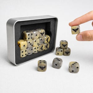 Puede incluir: Un juego de dados jaspeados en beige y gris con puntos negros y un emblema de pájaro negro en una cara. Los dados se guardan en una caja rectangular de metal plateado con un inserto de espuma negra, con varios dados sueltos fuera del estuche.