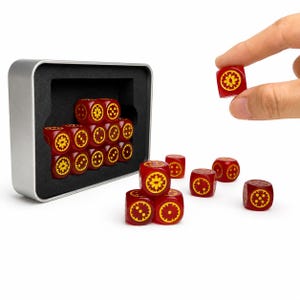 May include: A set of red six-sided dice with yellow circular gear-like symbols on each face. The dice are stored in a rectangular silver metal tin with a black foam insert. Several dice are arranged outside the tin, and one is held up.