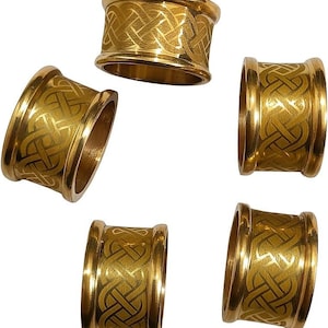 May include: A set of five gold-colored metal napkin rings, each featuring a continuous, engraved Celtic knot pattern around the exterior. The rings have a polished, reflective finish with raised edges and a cylindrical shape.