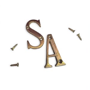 May include: Two metallic brass letters, S and A, each featuring two pre-drilled circular holes for mounting. Four small, matching brass-coloured screws are positioned around the letters, intended for securing the hardware to a surface.