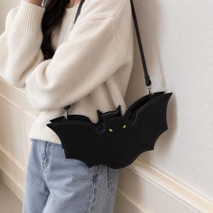 Può includere: Una borsa a tracolla nera a forma di pipistrello con finitura liscia, orecchie a punta e due piccoli occhi verde lime brillante. La borsa include una sottile tracolla nera fissata con hardware in metallo argentato, creando una distinta sagoma alata.