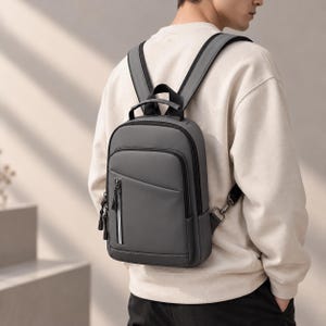 May include: A charcoal gray backpack featuring a smooth, matte finish, a vertical zippered front pocket with a white accent stripe, a top handle, and adjustable shoulder straps with black hardware, worn over a beige long-sleeve sweatshirt.