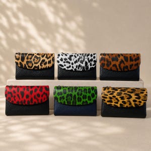 Pode incluir: Seis carteiras retangulares com abas dobráveis. A fila superior apresenta estampa de leopardo em marrom, branco e marrom. A fila inferior apresenta abas com estampa de leopardo vermelha, verde e amarela sobre corpos de couro texturizado.