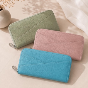 May include: Three rectangular zip-around wallets in sage green, dusty pink, and bright blue. Each wallet features a textured pebbled finish, a silver-toned metal zip pull, and a diagonal stitched seam detail across the front surface.