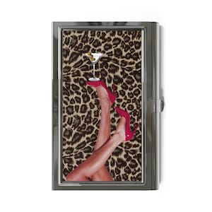 May include: A rectangular metal case featuring a leopard print background. The design displays a pair of legs wearing bright pink high-heeled pumps, with one foot balancing a martini glass containing a green olive on the heel.