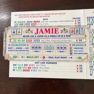 Pode incluir: Um suporte de cartão de Mah Jongg em acrílico transparente com o nome JAMIE em texto rosa, rodeado por instruções de jogo coloridas, ícones de naipes e regras de pontuação. A peça retangular inclui uma ranhura e detalhes florais.