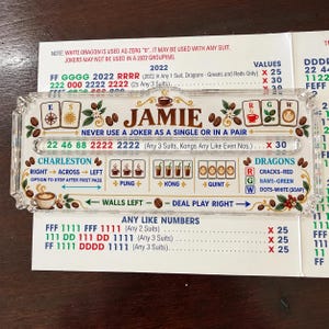 Peut inclure: Un porte-carte de Mah Jongg en acrylique transparent portant le nom JAMIE en lettres marron. La surface affiche des règles de jeu colorées, des icônes de tasses à café et des illustrations de grains de café, incluant le Charleston et les Dragons.
