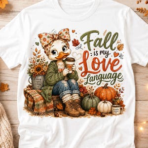 May include: A white t-shirt featuring a graphic of a cartoon duck wearing a floral bow, green cardigan, and boots, holding a coffee cup. The text reads Fall is my Love Language. The design includes pumpkins, autumn leaves, and a plaid blanket.