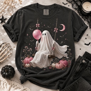 May include: A charcoal gray t-shirt featuring a graphic of a white ghost sitting on a swing. The ghost holds a pink balloon and is surrounded by pink pumpkins, small flowers, stars, bats, and a crescent moon. The design includes pink bows on the swing chains.