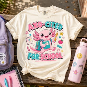 May include: A cream-colored short-sleeve t-shirt featuring a graphic of a pink axolotl carrying a teal backpack and a book. The text reads AXO-CITED FOR SCHOOL. Surrounding the axolotl are illustrations of a pencil, an apple, a ruler, and a notepad.