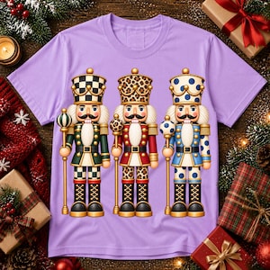 Puede incluir: Una camiseta de color morado claro con un estampado gráfico de tres figuras de cascanueces de pie una al lado de la otra. El cascanueces izquierdo tiene un patrón de cuadros verde y blanco, el central estampado de leopardo y el derecho lunares azules.