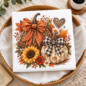 May include: A square white fabric print featuring two pumpkins. The larger pumpkin has a leopard print pattern and a large orange bow. The smaller pumpkin has a floral pattern and a black and white buffalo check bow. Includes a sunflower and heart.