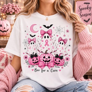 May include: A white t-shirt featuring a graphic print of three ghosts and three pumpkins in shades of pink. The ghosts wear pink bows and have pink ribbons. The pumpkins have pink plaid patterns and bows. Text reads Boo for a Cure.