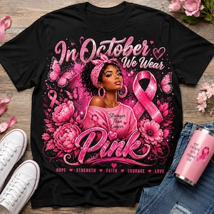 May include: A black t-shirt featuring a graphic of a person wearing a pink headscarf, surrounded by pink flowers, butterflies, and a pink ribbon. The text reads In October We Wear Pink, Stronger Than Cancer, Hope, Strength, Faith, Courage, Love.