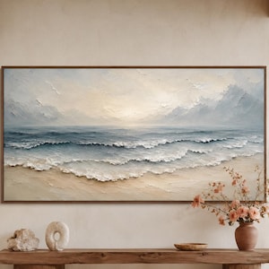 May include: A large rectangular landscape painting featuring a calm ocean scene with soft blue and white waves rolling onto a sandy beach under a pale sky. The artwork is displayed in a thin brown frame, showcasing textured brushstrokes.